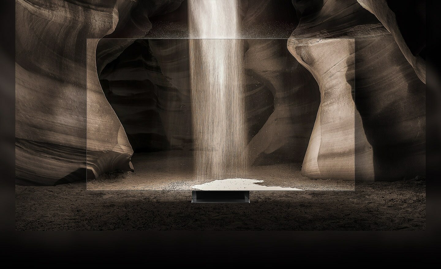 Telewizor QLED ustawiony w jaskini, z góry sypie się piasek. Krawędzie ekranu są tak wąskie, że wydaje się, jakby telewizora tam nie było.