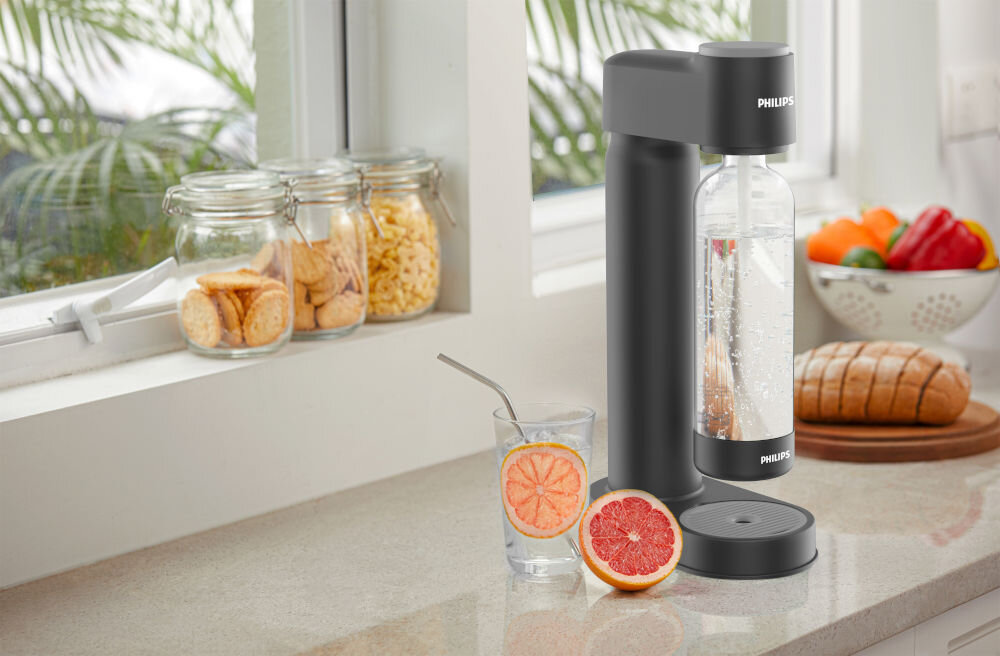 Na blacie kuchennym stoi czarny saturator PHILIPS z przezroczystą butelką wypełnioną wodą gazowaną. Obok znajduje się szklanka z plasterkiem grejpfruta, a w tle widać słoiki z przekąskami, chleb i misę z warzywami.