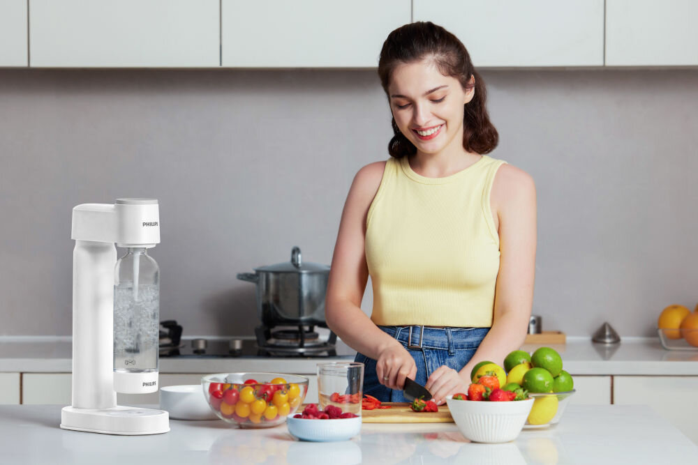 Kobieta kroi warzywa w kuchni, obok niej stoi saturator z logo PHILIPS. Na blacie znajdują się miski z owocami i warzywami oraz szklanka z napojem.