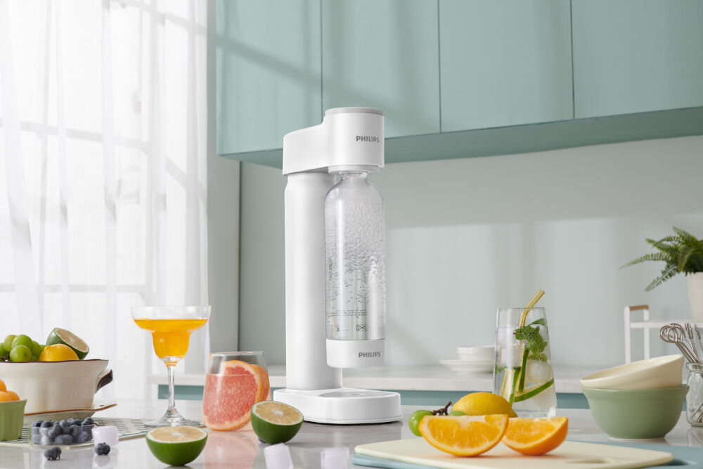 Na blacie kuchennym stoi biały saturator z przezroczystą butelką, obok której znajdują się owoce i szklanki z napojami. Na urządzeniu widoczne jest logo \'PHILIPS\'.
