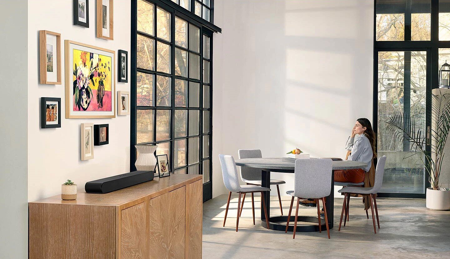 Kobieta siedzące przy stole w pokoju z szafką a na niej Soundbar S-seria HW-60B. HW-S60B/EN