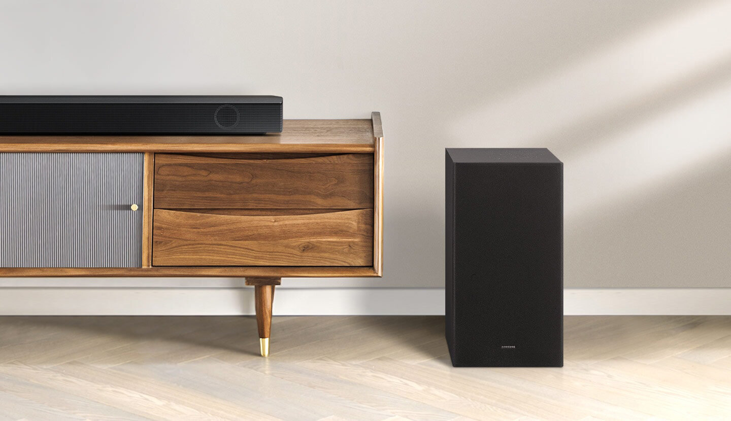 Soundbar Samsung serii B leży na szafce, obok szafki stoi pasujący do niego subwoofer. HW-B650/EN