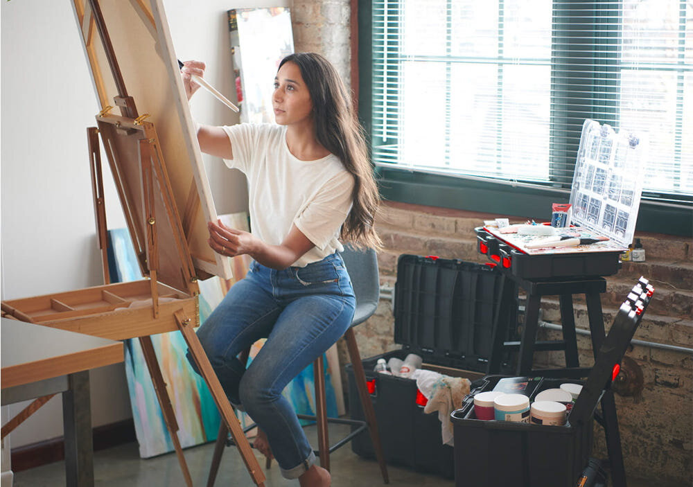 Organizer KETER 251491 osoba siedząca na stołku maluje na sztaludze w tle na stole pod oknem widoczne są czarne skrzynki i organizer z pokrywą i przegródkami metalowe zatrzaski z zapadką charakterystyczne „kliknięcie” waga około 2 kg wymiary 515 x 345 x 115 mm ergonomiczny uchwyt