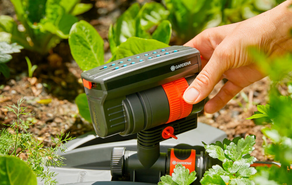 Zraszacz wahadłowy GARDENA Aquazoom Compact ogród rabata kwiatowa osoba trzyma dłoń na zraszaczu i pokrętłach prosty w montażu i obsłudze szybkie ustawienie uruchomienie systemu podlewania bez dodatkowych narzędzi samodzielne dopasowanie parametrów podlewania