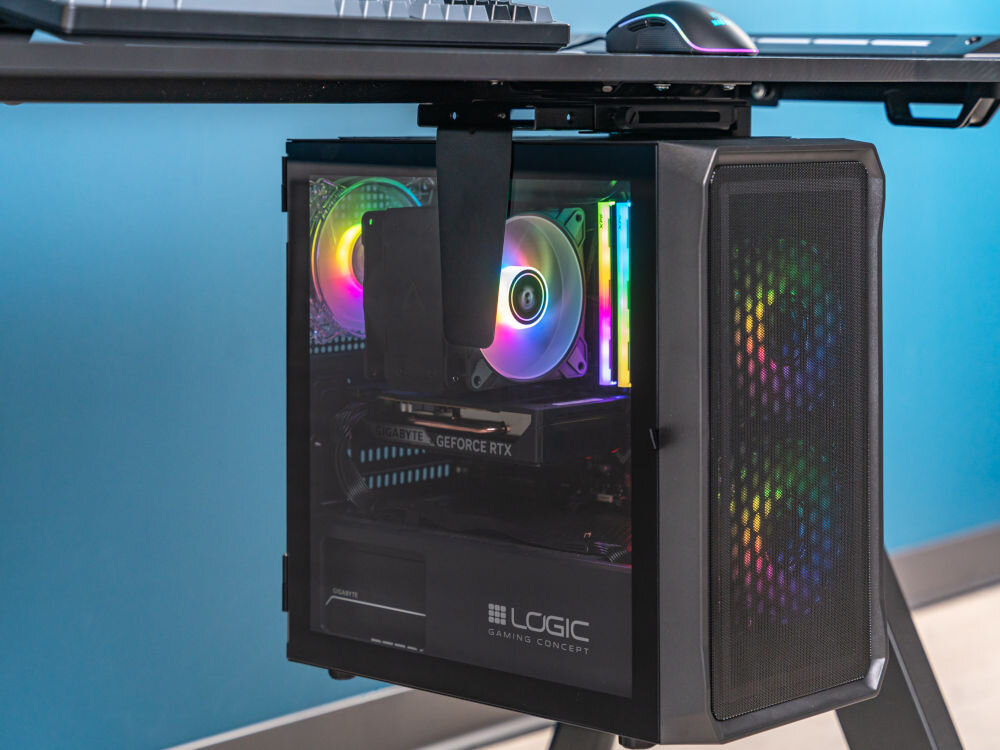 Uchwyt biurkowy do obudowy PC ART M-01 Komputer stacjonarny gamingowy z podświetleniem RGB zawieszony pod blatem biurka za pomocą czarnego, regulowanego uchwytu biurkowego. Pokazuje, jak uchwyt oszczędza miejsce na biurku.