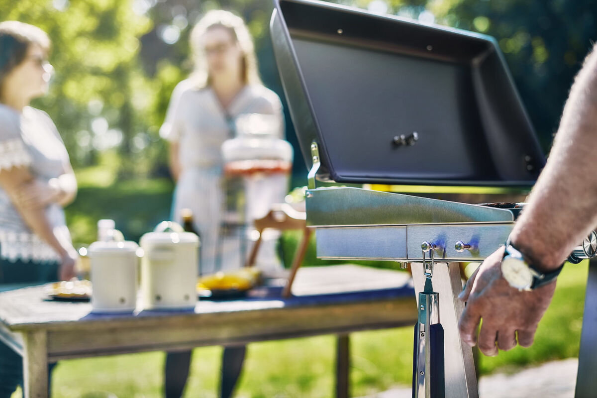 Grill elektryczny KALORIK TKG EBBQ 1002 L, elementy, pokrywa, dotyk, podstawa, w tle kobiety rozmawiają, na pierwszym planie osoba pracuje przy grillu w ogrodzie