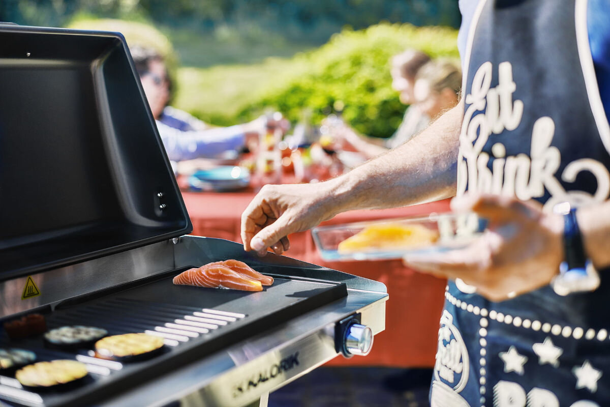 Grill elektryczny KALORIK TKG EBBQ 1002 L, niezależne, dwa, odmienne, strefy, osoba obsługuje 1 z platform grilla, w tle ogród