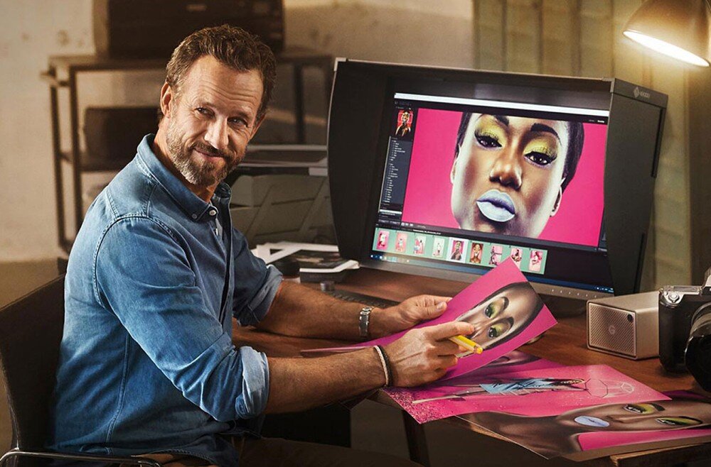 Monitor EIZO ColorEdge CG2700S Mężczyzna w dżinsowej koszuli siedzi przy biurku, trzyma w dłoni wydrukowaną grafikę kobiety o intensywnym makijażu. Na monitorze przed nim widoczny jest ten sam portret w programie do edycji zdjęć, opis