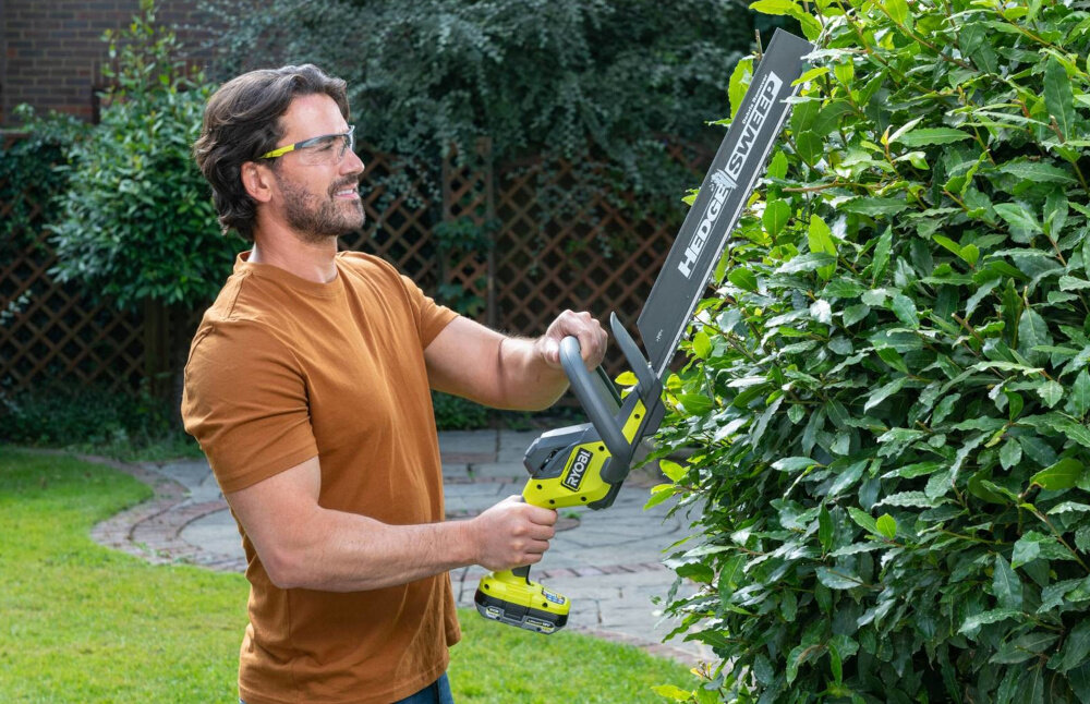 Nożyce do żywopłotu RYOBI RY18HT50A-0 500mm 18V akumulatorowe ogród osoba stoi przycina żywopłot w tle drzewa krzewy silnik szczotkowy stabilna i przewidywalna praca wspiera równomierne przenoszenie mocy na ostrze płynność ruchów ogranicza nagłe spadki wydajności