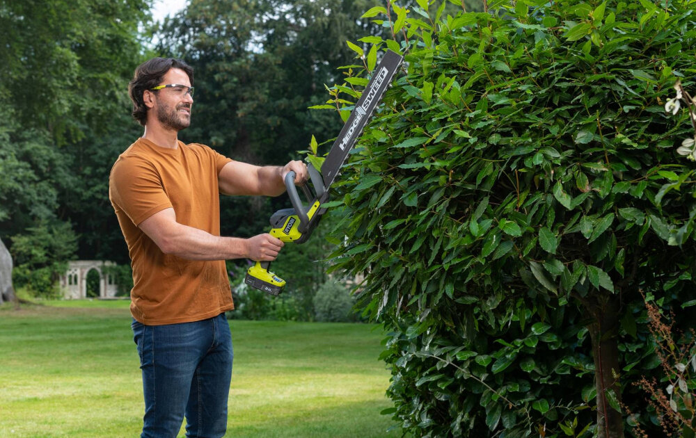 Nożyce do żywopłotu RYOBI RY18HT50A-0 500mm 18V akumulatorowe ogród osoba przycina żywopłot w tle drzewa krzewy długość ostrza 50 cm maksymalna zdolność cięcia do 24 mm większa efektywność działań równomierne przycięcie na całej długości roślin