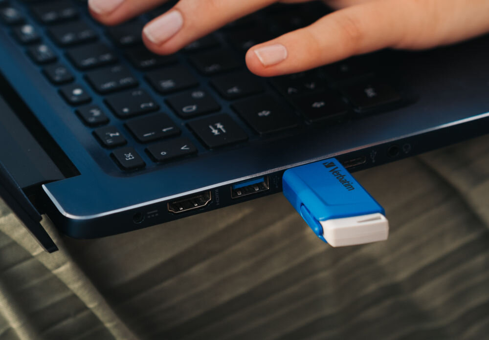 Na zdjęciu widać niebieski pendrive marki Verbatim podłączony do laptopa. Obok znajduje się dłoń z palcami na klawiaturze.