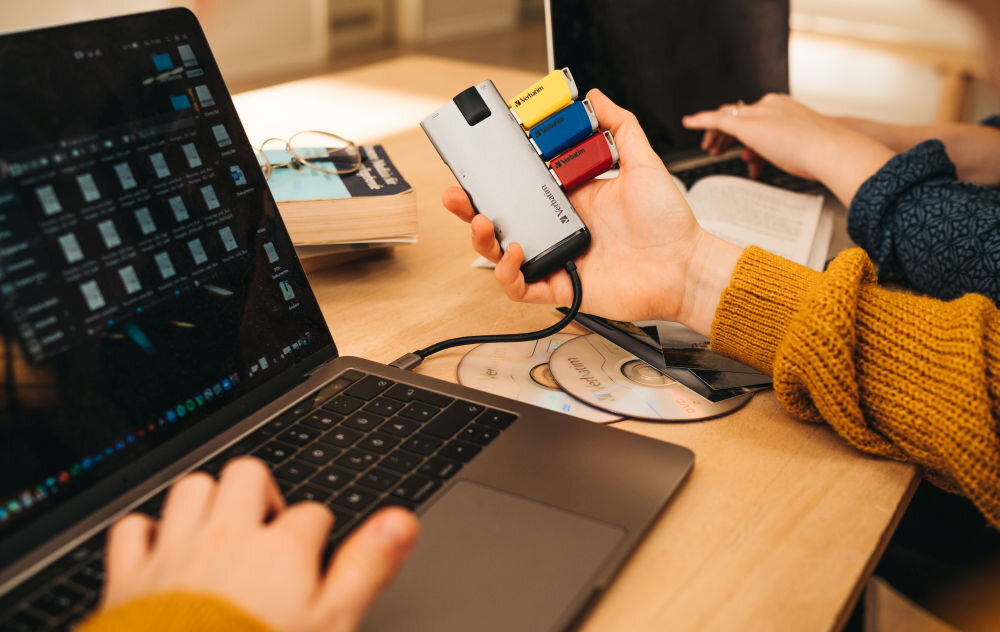 Osoba trzyma w ręku urządzenie z trzema pendrive\'ami w kolorach czerwonym, niebieskim i żółtym, podłączone do laptopa. Na stole leżą książki i płyty CD.