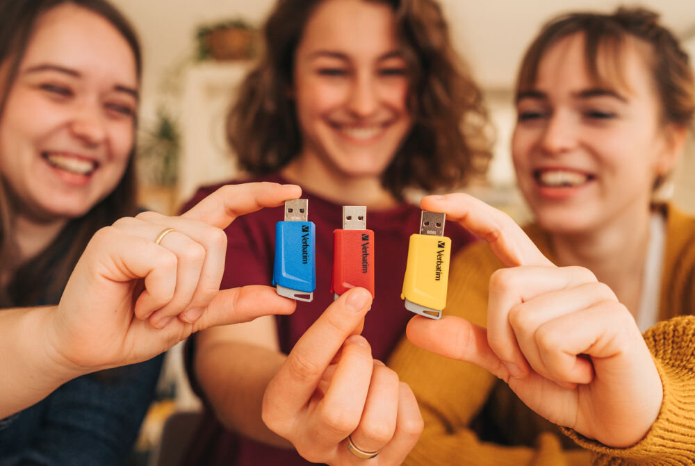 Trzy osoby trzymają w dłoniach pendrive\'y w kolorach niebieskim, czerwonym i żółtym z logo \'Verbatim\'.