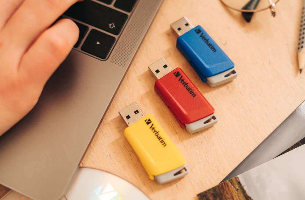 Na drewnianym biurku obok laptopa leżą trzy pendrive\'y w kolorach żółtym, czerwonym i niebieskim z logo \'Verbatim\'.