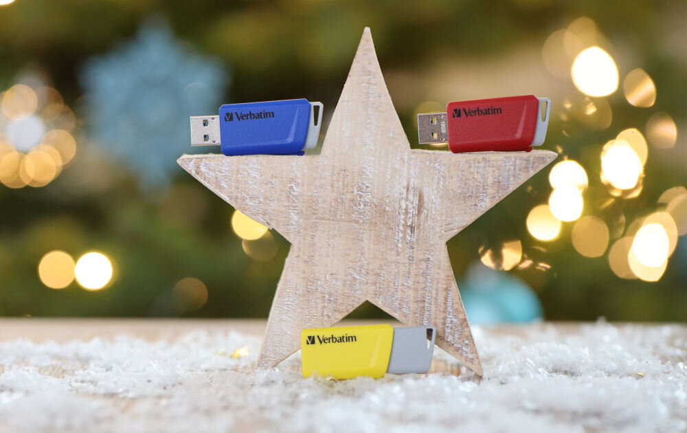 Trzy pendrive\'y Verbatim w kolorach niebieskim, czerwonym i żółtym są umieszczone na drewnianej gwieździe. W tle widoczne są rozmyte światełka, przypominające dekoracje świąteczne.