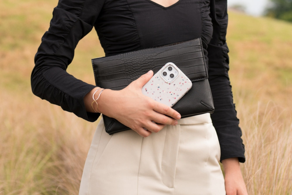 Osoba trzyma czarne etui i telefon w różowym etui z kolorowymi plamkami. W tle widoczne jest pole trawy.