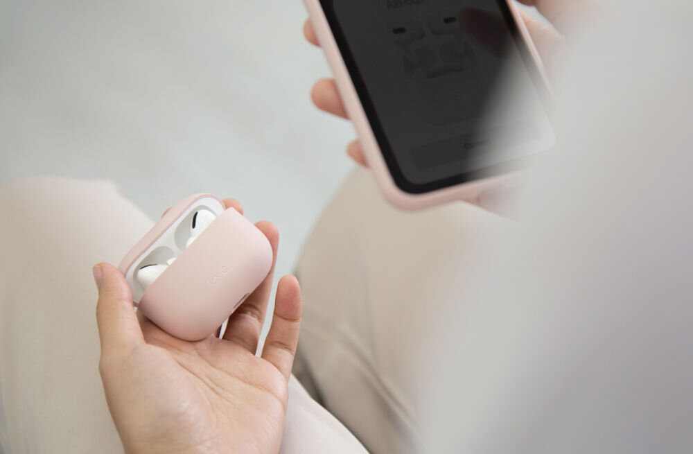 Osoba trzyma różowe etui na słuchawki, w którym znajdują się białe słuchawki. W drugiej ręce trzyma smartfon z wyświetlonym ekranem.