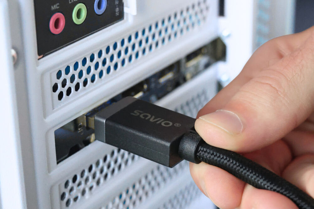 Kabel DisplayPort - DisplayPort SAVIO CL-166 2 m Ręka podłącza wtyczkę kabla DisplayPort marki SAVIO z czarnym, plecionym oplotem do odpowiedniego portu w tylnej części białej obudowy komputera stacjonarnego (wieży).