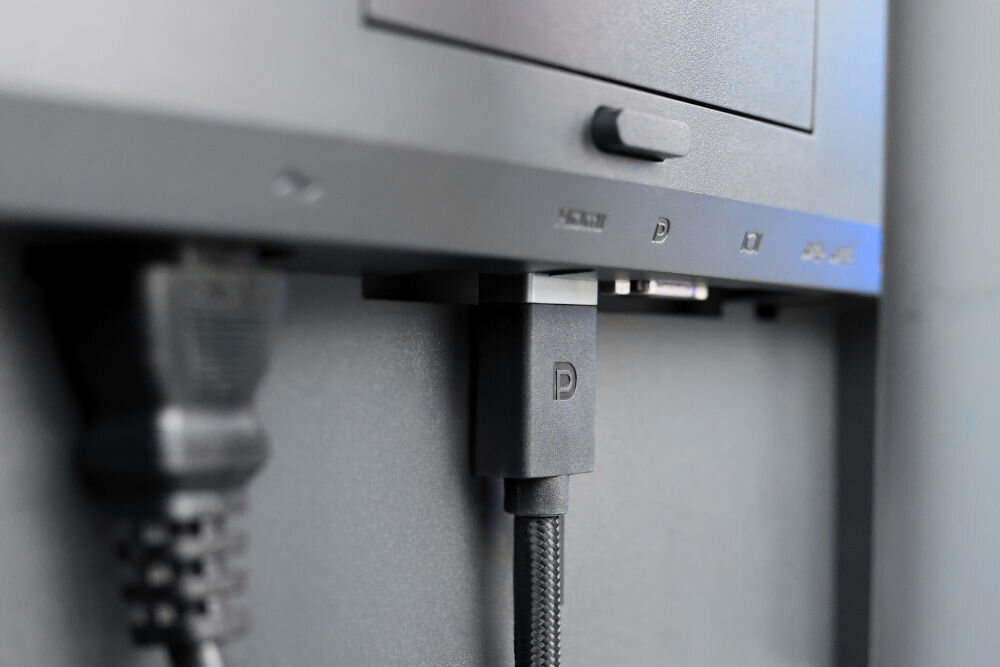 Kabel DisplayPort - DisplayPort SAVIO CL-166 2 m Zbliżenie na porty w tylnej części monitora. Do portu DisplayPort (oznaczonego literą 'D') podłączona jest czarna wtyczka kabla o plecionym oplocie. Obok widoczna jest również podłączona wtyczka zasilania lub innego kabla.