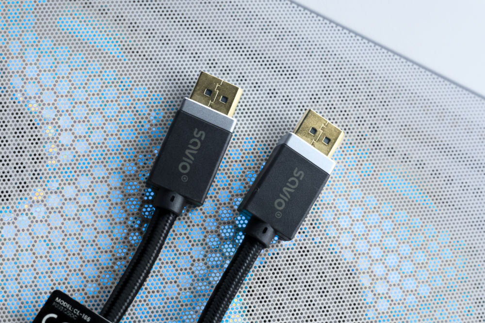 Kabel DisplayPort - DisplayPort SAVIO CL-166 2 m Dwie wtyczki kabla DisplayPort marki SAVIO leżą na powierzchni przypominającej metalową kratkę lub siatkę wentylacyjną komputera, która delikatnie podświetla się na niebiesko. Wtyczki mają pozłacane końcówki i aluminiowo-szare akcenty.