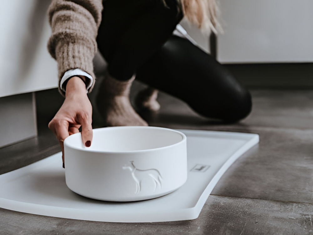 Miska ceramiczna dla psa HUNTER Osby 68982 Antracytowy, wysokiej jakości ceramika, elegancki design z tłoczonym logo, ozdoba wnętrza - osoba stawia na podłodze białą, ceramiczną miskę dla psa z tłoczonym logo na jasnej, antypoślizgowej podkładce