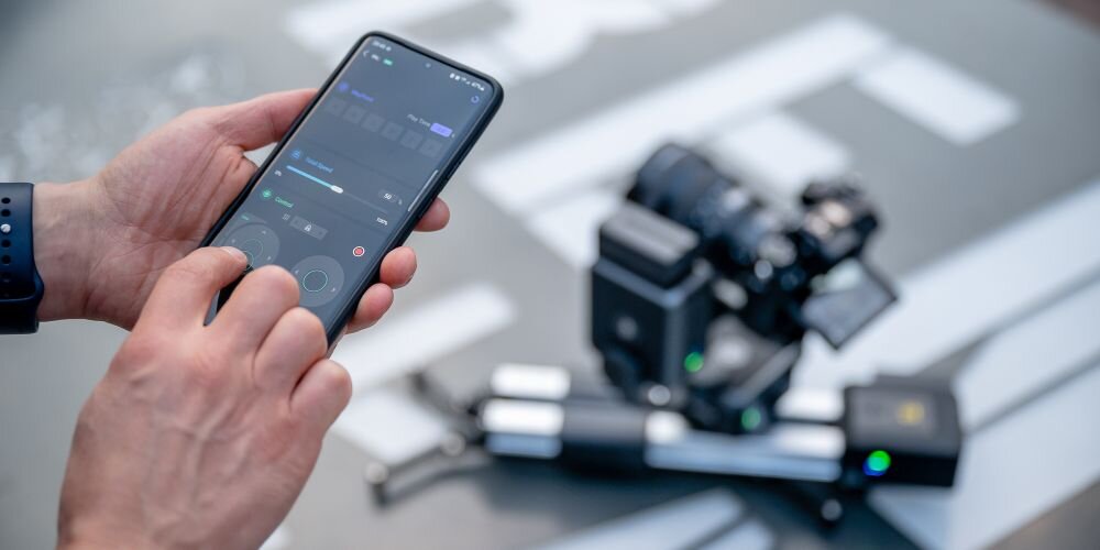 Męskie dłonie obsługują aplikację Zeapon Lab na telefonie. W tle rozmyty sprzęt na sliderze Zeapon sterowany z tegoż telefonu