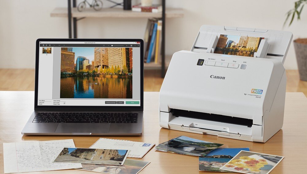 Na stole znajduje się laptop z wyświetlonym zdjęciem budynków oraz skaner Canon imageFORMULA RS40, który skanuje zdjęcia. Obok leżą różne fotografie i kartki z tekstem.
