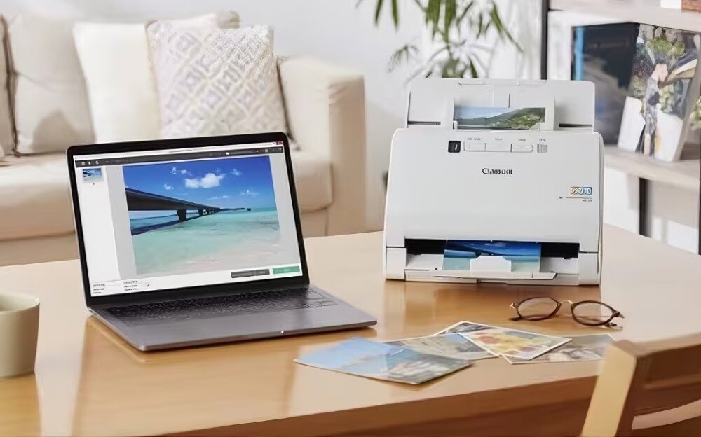 Na drewnianym stole znajduje się otwarty laptop z wyświetlonym zdjęciem plaży oraz skaner z logo Canon, w którym widać włożone zdjęcia. Obok leżą okulary i kilka fotografii.