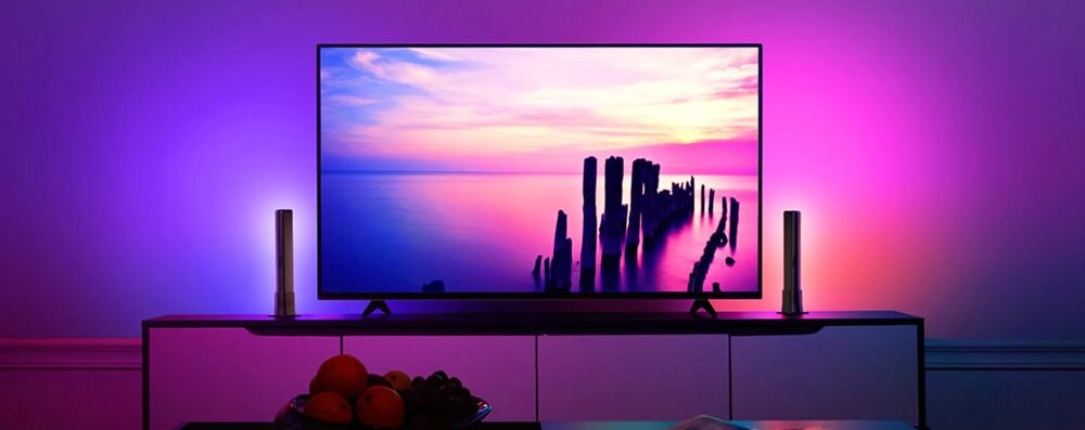 Lampa GOVEE H6056 Flow Plus RGBICWW Wi-Fi, światło, kolor, wnętrze, styl, Duży TV z podświetleniem Govee Glide po bokach, pokazujący zachód słońca nad morzem, z purpurowymi i różowymi efektami LED rozświetlającymi pokój i owoce na stoliku.