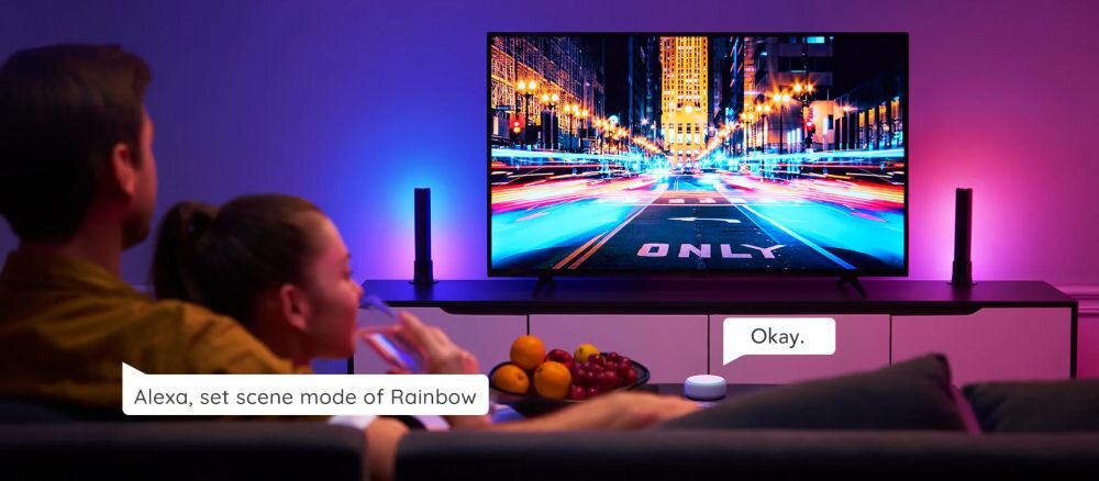 Lampa GOVEE H6056 Flow Plus RGBICWW Wi-Fi, muzyka, rytm, reakcja, dźwięk, Para na sofie ogląda miasto nocą na dużym TV z synchronizowanym oświetleniem Govee Glide po bokach, sterowanym głosowo przez Alexę: „Set scene of rainbow”, z owocami na stoliku.