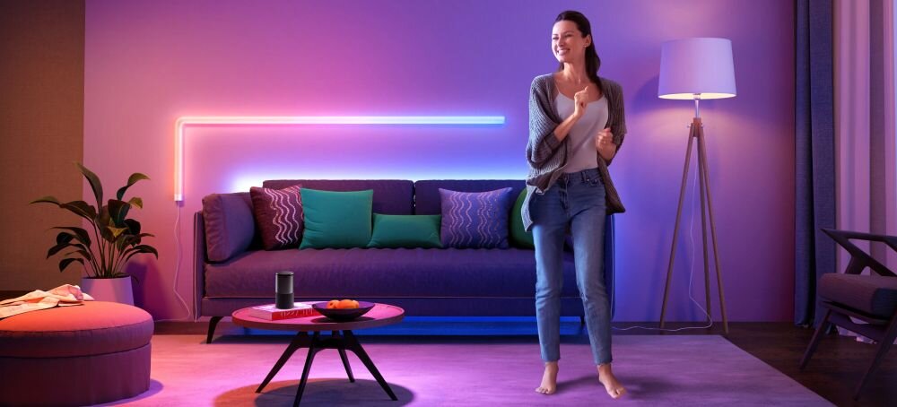 Lampa GOVEE H6062 Glide Wi-Fi/Bluetooth (7 szt.), kolor, światło, styl, klimat, Młoda kobieta w luźnym swetrze steruje neonową listwą LED Govee na ścianie salonu za fioletową sofą, z lampą podłogową i stolikiem w tle.