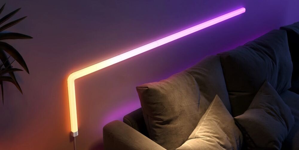 Lampa GOVEE H6062 Glide Wi-Fi/Bluetooth (7 szt.), muzyka, rytm, gra, impreza, Elastyczna neonowa taśma LED Govee w kształcie litery L na ścianie salonu, świecąca gradientem pomarańczowo-fioletowym, obok szarej sofy i rośliny doniczkowej.
