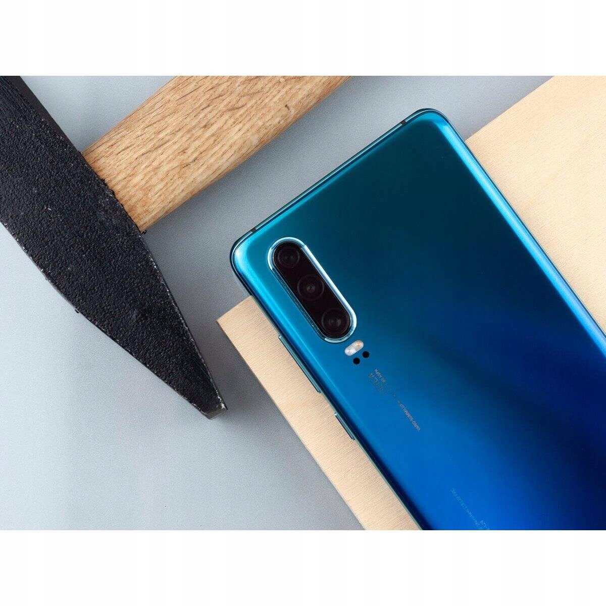 Na obrazie widać tylną część niebieskiego smartfona z trzema obiektywami aparatu. Obok telefonu znajduje się drewniany młotek z czarną główką.