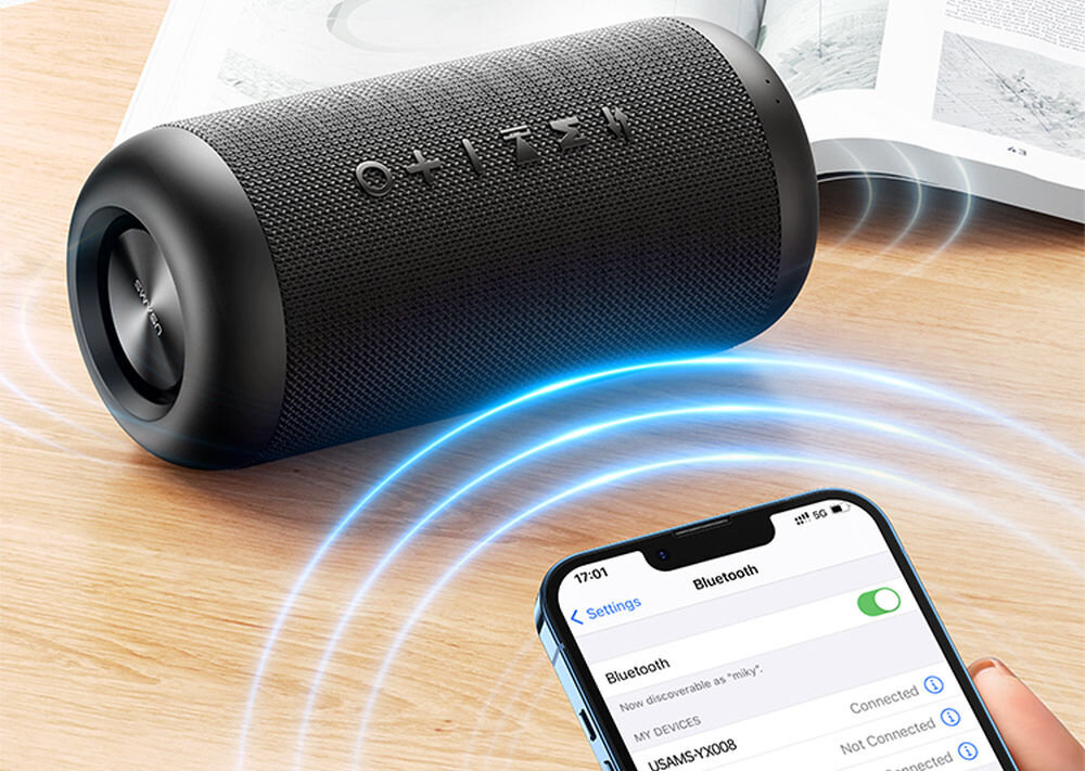 Czarny głośnik mobilny o cylindrycznym kształcie leży na drewnianej powierzchni, obok znajduje się smartfon z włączonym Bluetooth i widoczną nazwą urządzenia \'USAMS-YX008\'.