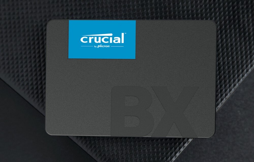 Dysk CRUCIAL BX500 Czarny dysk SSD z niebieską naklejką leży na ciemnym, teksturowanym tle, technologia, konstrukcja, testy jakości