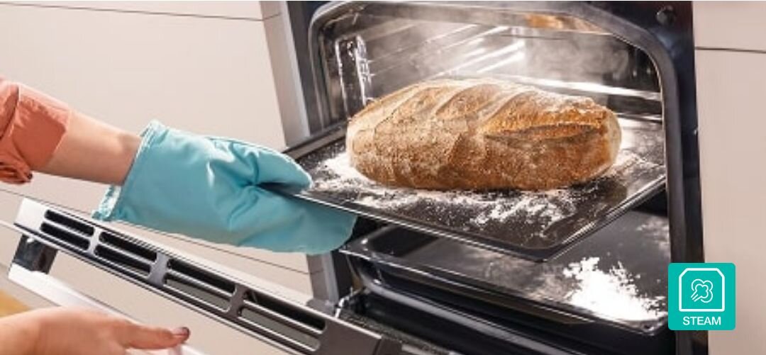 KUCHNIA GORENJE GECS5C70XPA Osoba w turkusowej rękawicy kuchennej wysuwa z piekarnika blachę z dużym, parującym bochenkiem chleba, a unosząca się para i ikona „STEAM” w rogu podkreślają funkcję pieczenia z dodatkiem pary pieczenie z parą wodną