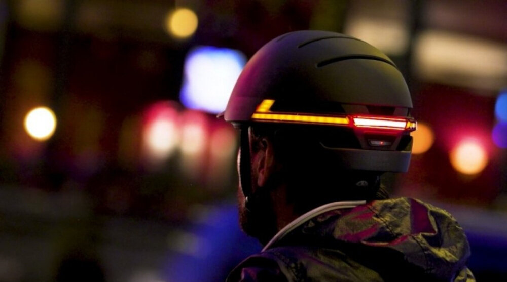 Osoba nosi kask z wbudowanym oświetleniem LED, które świeci na czerwono. Tło jest rozmyte i kolorowe, co sugeruje miejskie otoczenie nocą.