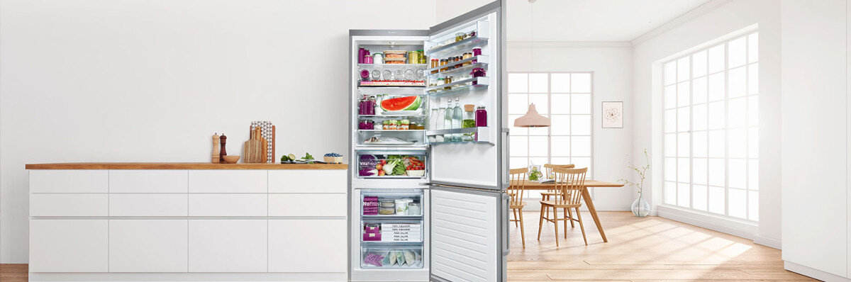 LODÓWKA BOSCH KGN49LBCF BLACK Zdjęcie przedstawia otwartą lodówkę stojącą w jasnej, nowoczesnej kuchni, wypełnioną świeżymi produktami rozłożonymi na półkach i w szufladach. W tle widać przestronne pomieszczenie z dużymi oknami i drewnianym stołem powiększona przestrzeń szerokość pojemność