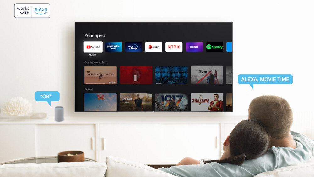 Na obrazie widać telewizor z interfejsem Google TV, na którym wyświetlane są aplikacje takie jak YouTube, Netflix i Spotify. Obok telewizora znajduje się głośnik z napisem \'OK\', a w prawym górnym rogu widnieje logo \'works with alexa\'.