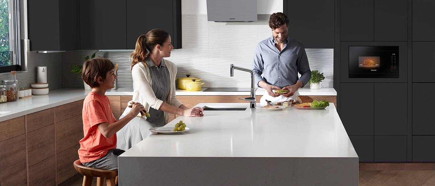 Rodzina zgromadzona przy kuchennej wyspie, a w tle działająca mikrofala Samsung MG22T8254AB – model dostępny w sklepach Media Expert Rodzina zgromadzona przy kuchennej wyspie, a w tle działająca mikrofala Samsung MG22T8254AB – model dostępny w sklepach Media Expert