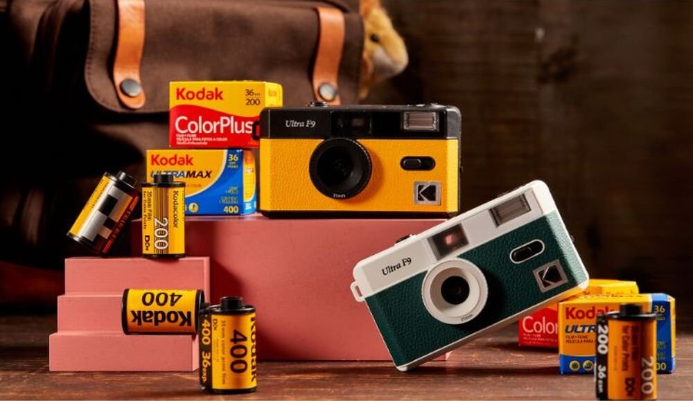 Na obrazie znajdują się dwa aparaty analogowe KODAK Ultra F9 w kolorach żółtym i zielonym, otoczone opakowaniami filmów fotograficznych Kodak. W tle widoczna jest brązowa torba.