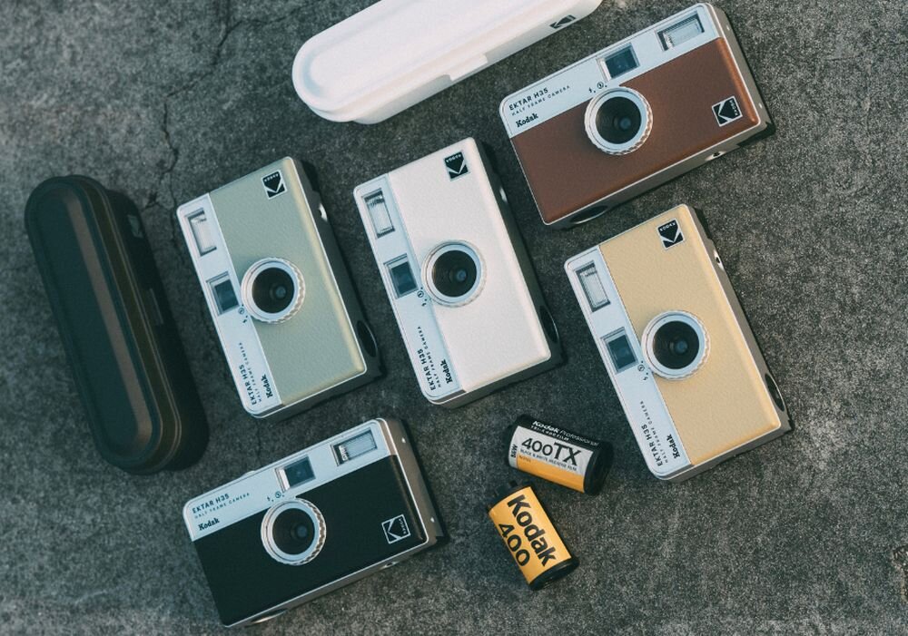 Na obrazie widoczne są cztery aparaty analogowe w różnych kolorach oraz dwa pojemniki na film fotograficzny z napisami \'400TX\' i \'Kodak 400\'. Aparaty mają logo Kodak i są ułożone na szarej powierzchni.