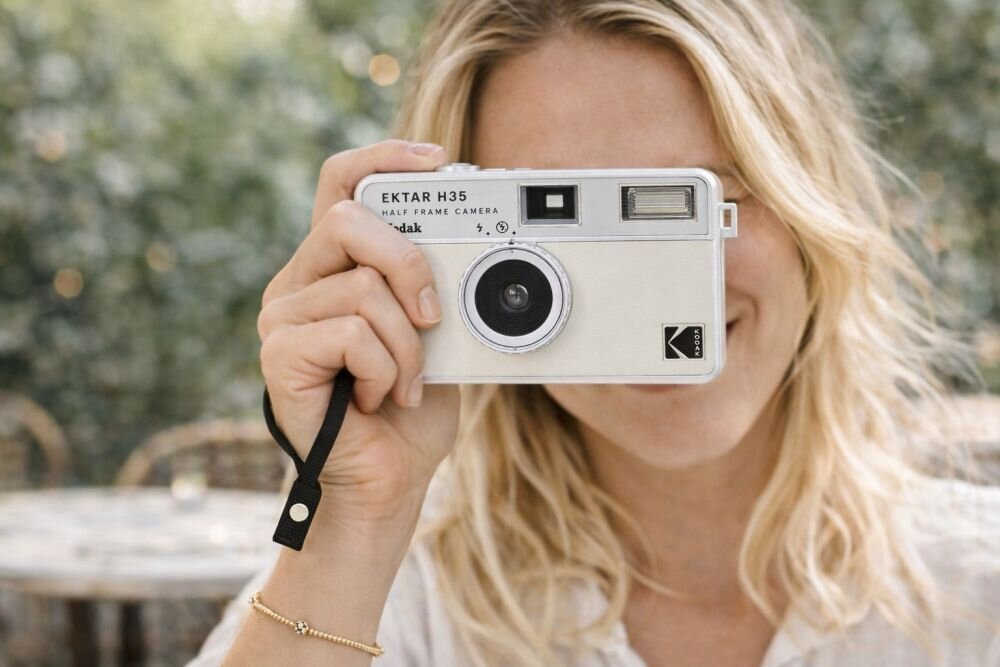 Osoba trzyma aparat analogowy z widocznym napisem \'EKTAR H35 HALF FRAME CAMERA Kodak\'. Na aparacie znajduje się logo \'Kodak\'.