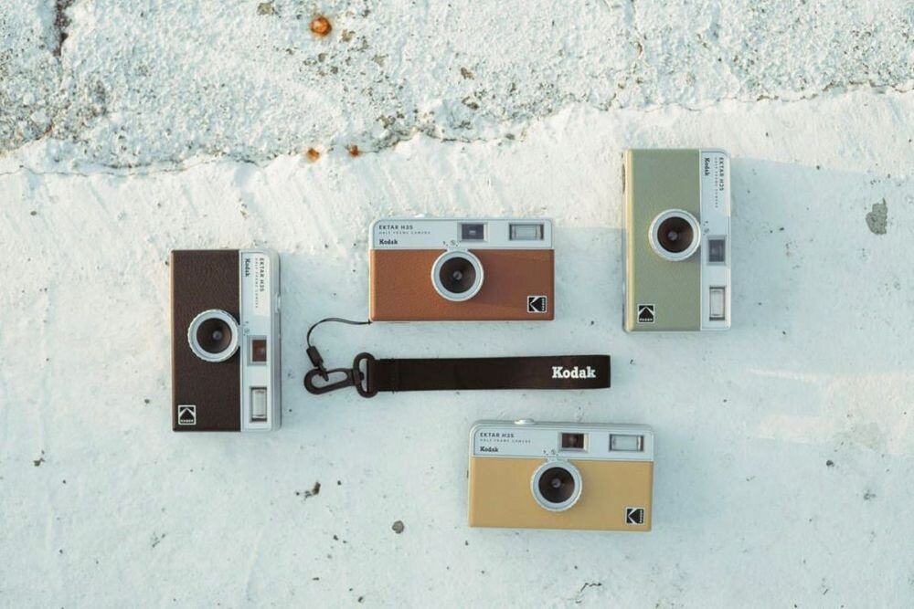 Na obrazie widoczne są cztery aparaty analogowe KODAK Ektar H35 w różnych kolorach: czarnym, brązowym, zielonym i beżowym. Obok aparatów leży czarny pasek z napisem \'Kodak\'.