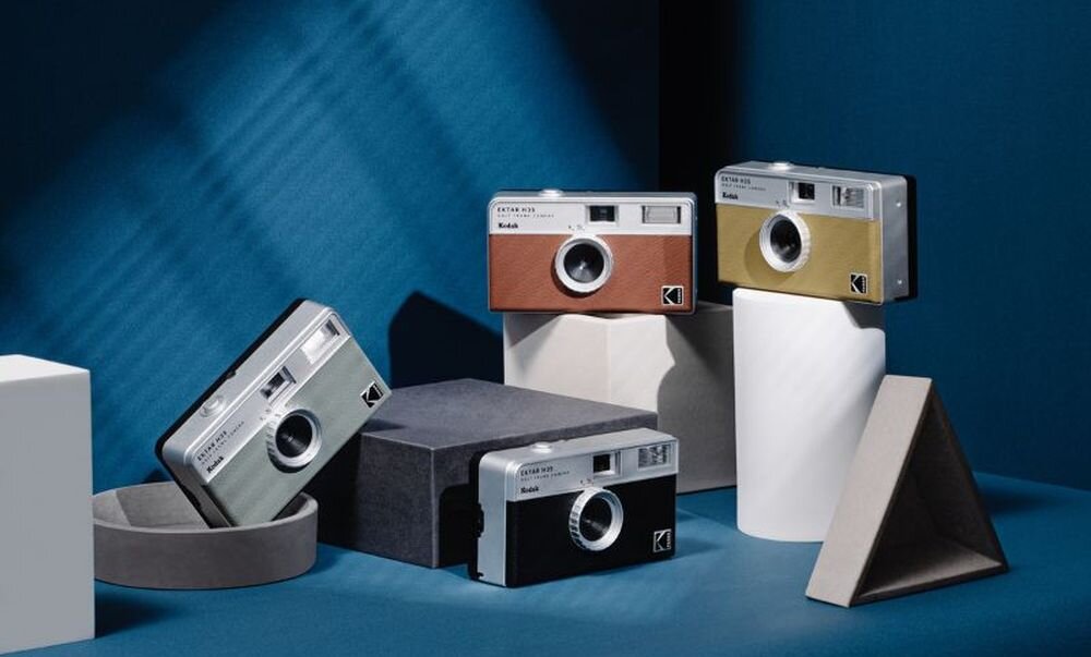 Na obrazie znajdują się cztery aparaty analogowe w różnych kolorach: czarnym, brązowym, złotym i srebrnym, ustawione na geometrycznych kształtach. Na każdym aparacie widnieje napis \'Kodak\'.