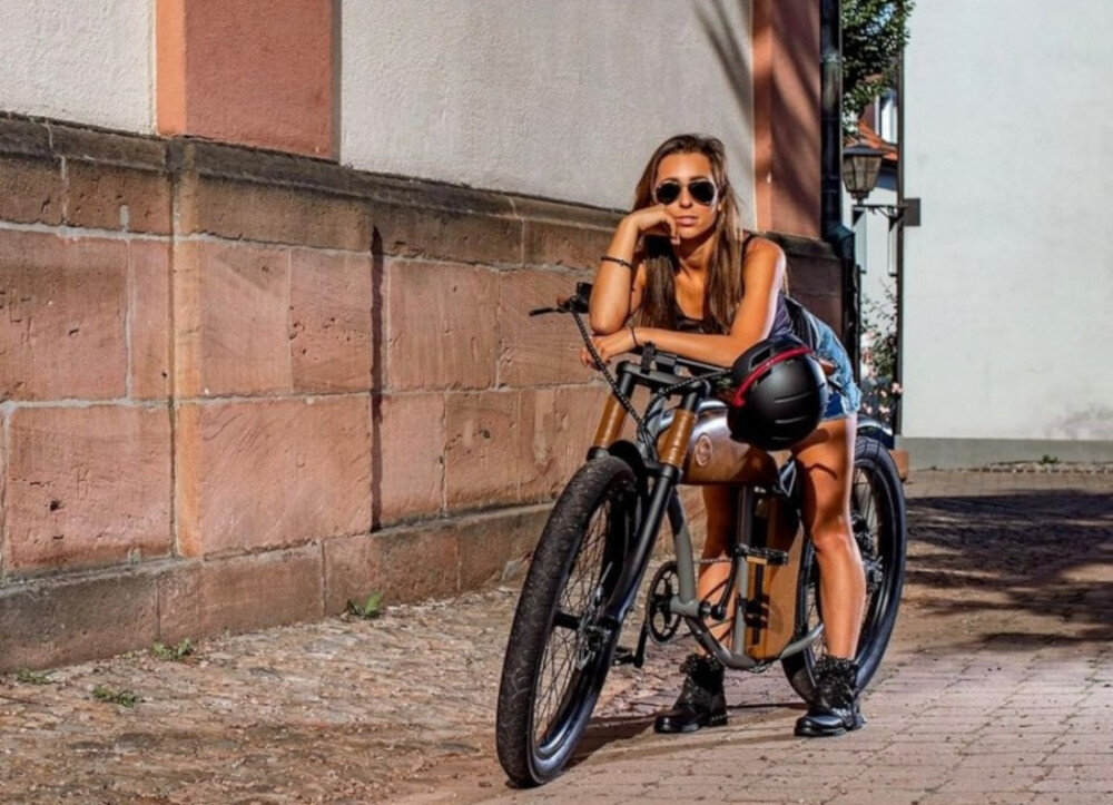 Osoba siedzi na rowerze opartym o ścianę z cegieł, trzymając kask rowerowy w kolorze szarym z lampką. W tle widać fragmenty budynków i latarnię uliczną.