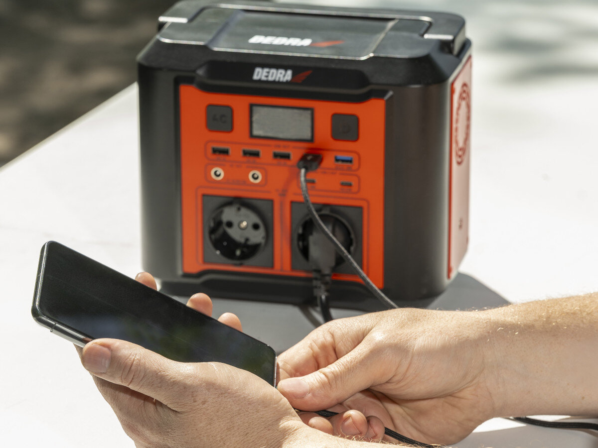 Stacja zasilania DEDRA DEZS0300 296Wh Bank energii porty gniazda wejścia oświetlenie, na stole składanym stoi stacja, na pierwszym planie ładowanie smartfona przez stacje