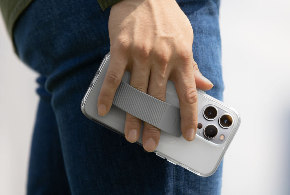 Osoba trzyma przezroczyste etui na telefon z paskiem, które jest założone na smartfonie z trzema obiektywami aparatu. Etui ma widoczny wzór na pasku, a w tle widać fragment ubrania z dżinsu.