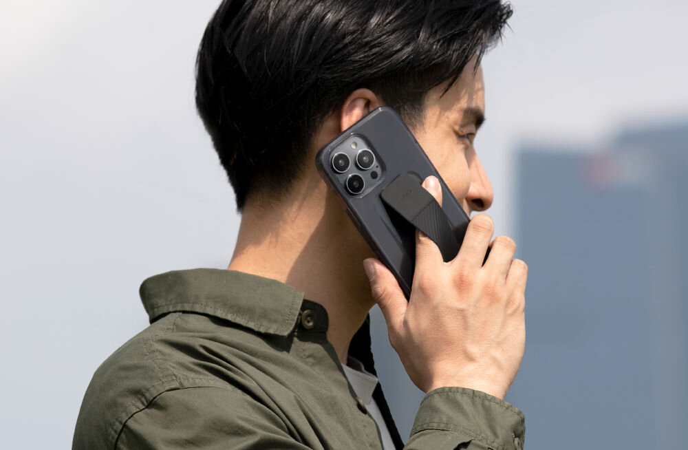 Osoba trzyma przy uchu telefon z szarym etui, które ma widoczny uchwyt. W tle widać rozmyty krajobraz miejski.