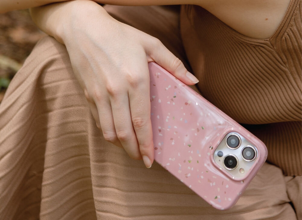 Osoba trzyma różowe etui z wzorem terrazzo na telefonie z trzema aparatami. Etui ma delikatne, kolorowe plamki na powierzchni.